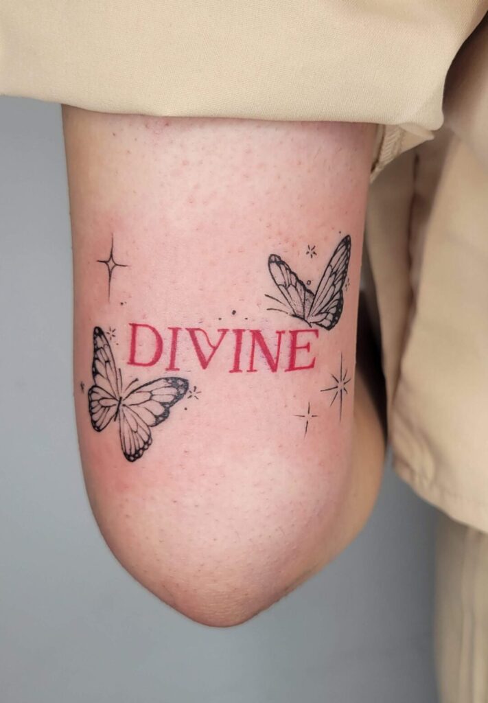 Fine line butterfly tattoo with red “DIVINE” lettering on the elbow, minimalist black butterflies with small stars, delicate feminine tattoo design.