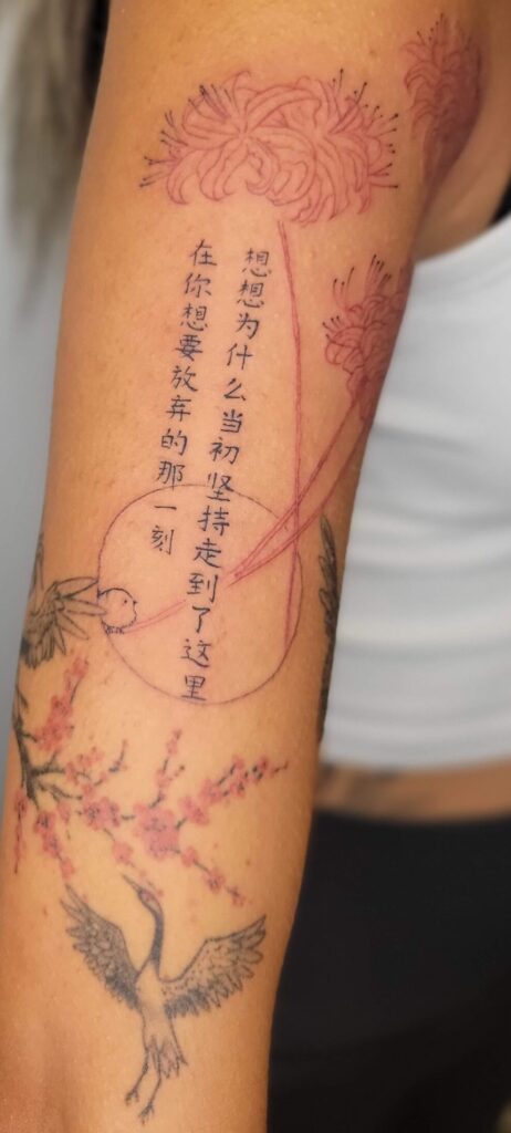 Fine line script tattoo with Chinese lettering and floral linework on upper arm, minimalist vertical text tattoo design in Canada.