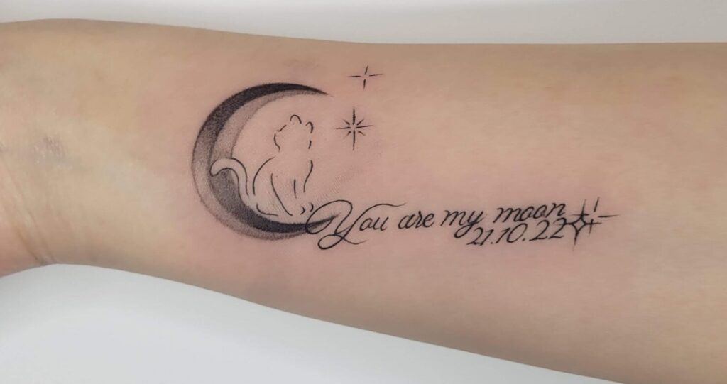 Minimal fine line tattoo of a cat sitting on crescent moon with stars and small script lettering on forearm, delicate minimalist tattoo design in Calgary